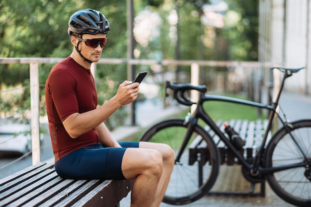 Ciclista sportivo in casco e occhiali che utilizza un moderno telefono cellulare mentre è seduto su una panca di legno vicino alla bici nera Sportivo che si prende una pausa durante l'allenamento all'aperto