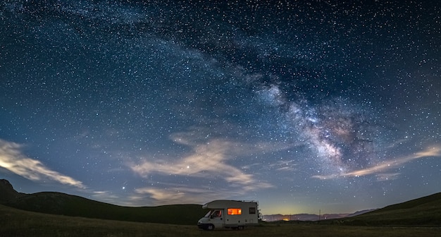 Cielo notturno panoramico sugli altopiani di Campo Imperatore, Abruzzo, Italia. L'arco e le stelle della galassia della Via Lattea sopra il camper illuminato. Libertà di campeggio in un paesaggio collinare unico.