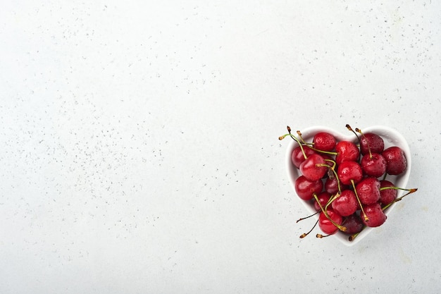 Ciliegia con gocce d'acqua sul piatto a forma di cuore su tavola di pietra bianca. Ciliegie mature fresche. Ciliegie rosse dolci. Vista dall'alto. Stile rustico. Sfondo di frutta