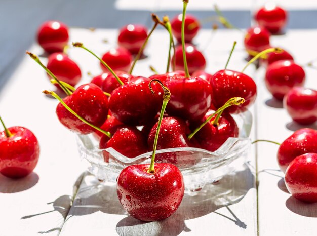 Ciliegia dolce in una ciotola di vetro e sparsa su un tavolo di legno bianco Le gocce di ciliegia fresca rossa lavate brillano