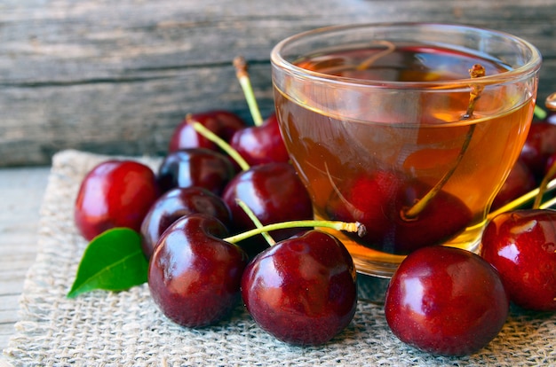 Ciliegie mature e tè appena raccolti in una tazza di vetro trasparente su un panno di tela. Tè di ghiaccio della frutta e frutti della ciliegia sulla vecchia tavola di legno. Concetto di bevanda estiva.