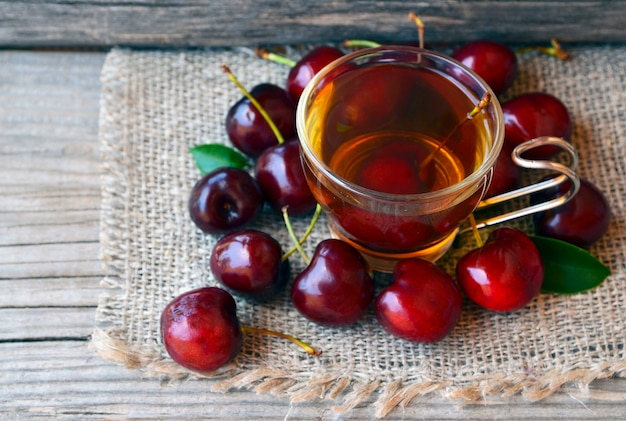Ciliegie mature e tè appena raccolti in una tazza di vetro trasparente su un panno di tela. Tè di ghiaccio della frutta e frutti della ciliegia sulla vecchia tavola di legno. Concetto di bevanda estiva.