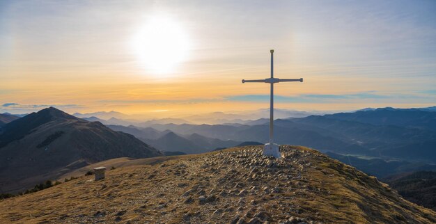 Cima della montagna con una grande croce bianca con montagne sullo sfondo e sole con cielo arancione