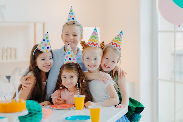 Cinque simpatici bambini indossano cappucci a cono festivo che si abbracciano e fanno foto insieme, giocano e festeggiano il compleanno hanno espressioni felici in posa al tavolo festivo