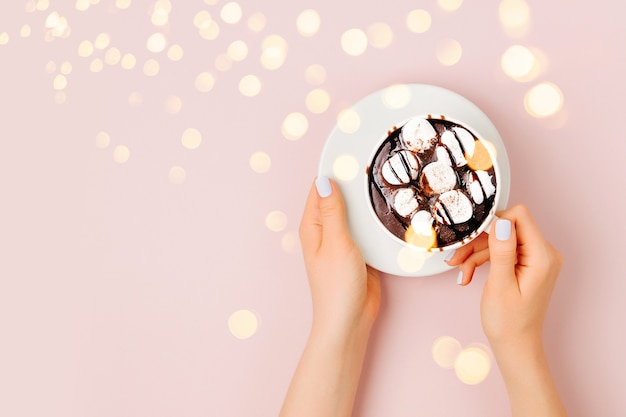 Cioccolata calda con marshmallow in mano di donna su sfondo rosa pallido