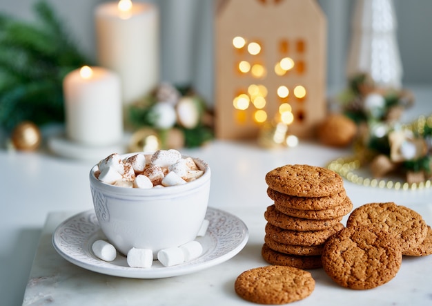 Cioccolata calda con marshmallow in tazza bianca circondata da decorazioni natalizie invernali su tavolo bianco