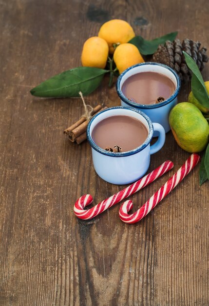 Cioccolata calda in tazze di metallo smaltato, mandarini freschi, bastoncini di cannella, pigna e bastoncini di zucchero sul tavolo di legno rustico