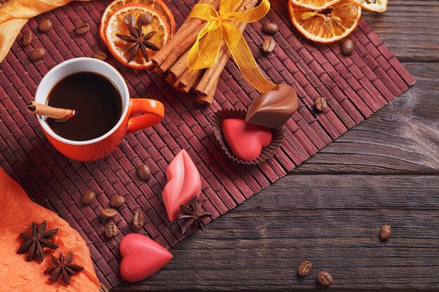 Cioccolatini a forma di cuore e labbra, tazza arancione di caffè nero, chicchi di caffè tostati neri, fette di limone essiccate con spezie, cannella e anice su sfondo scuro. Concetto di dessert al cioccolato