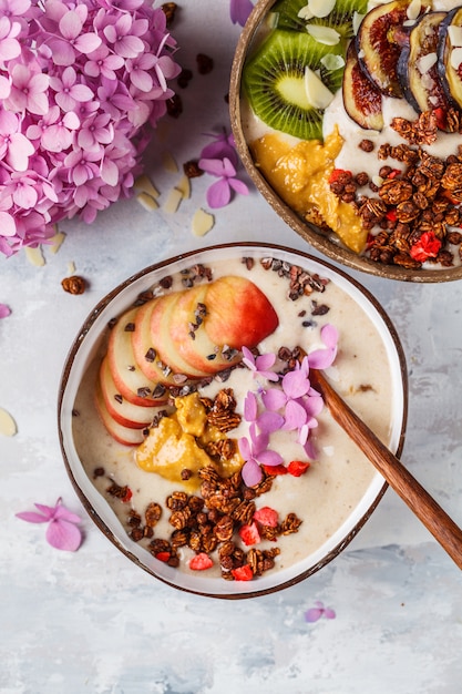 Ciotola del frullato con frutta e granola con la ciotola delle coperture della noce di cocco su fondo grigio. Cibo vegano sano.