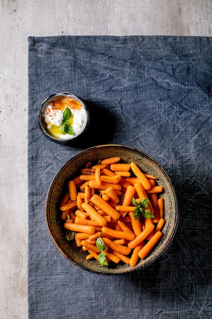 Ciotola di carota intera al forno con salsa di yogurt sulla tovaglia blu su sfondo grigio. Piatto vegano. Disposizione piatta, copia spazio