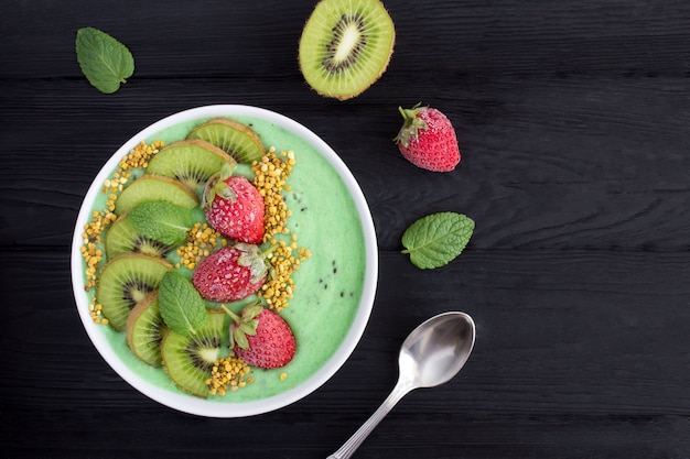 Ciotola di frullati con banana, kiwi, fragola congelata e polline di ape sullo sfondo di legno nero. Vista dall'alto. Copi lo spazio.