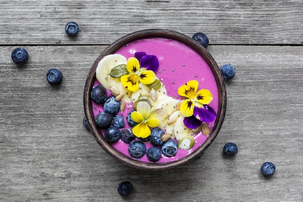 Ciotola di frullato di mirtilli condita con bacche fresche, banana, semi di chia e fiori per una sana colazione vegetariana.