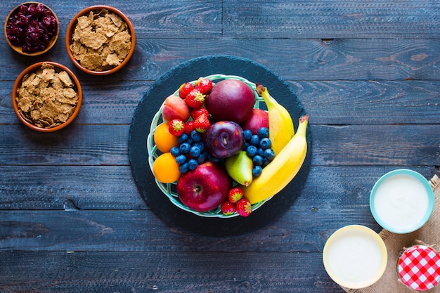 Ciotola di frutta fresca con banana, mela, fragole, albicocche, mirtilli, prugne, cereali integrali, forchette, vista dall'alto