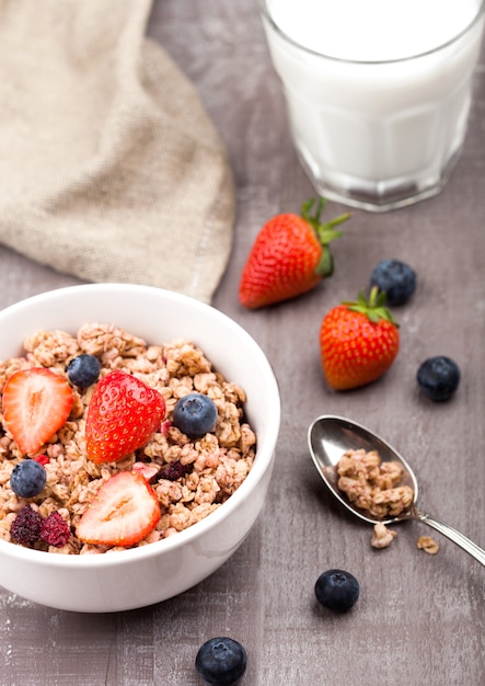 Ciotola di muesli sano del cereale con le fragole e mirtilli e bicchiere di latte sul bordo di legno