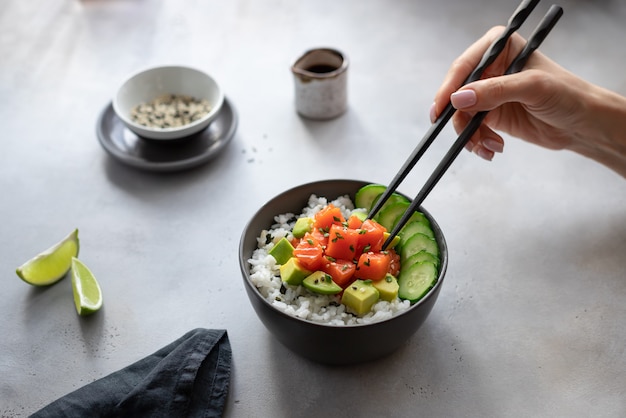 Ciotola di poke hawaiana delle bacchette di cibo della mano della donna con i salmoni, il riso, l'avocado ed il cetriolo. Dieta alimentare.