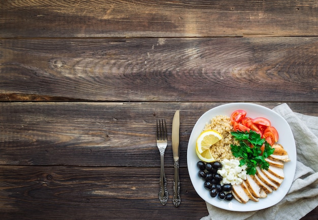 Ciotola di quinoa pollo sano con pomodorini, feta, olive e prezzemolo su superficie di legno rustica. Vista dall'alto con spazio per il testo.