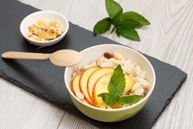 Ciotola in porcellana con porridge di sorgo con pesca fresca, anacardi e mandorle, cucchiaio di legno e ciotola con noci su tavola di pietra. Insalata di sorgo vegana senza glutine con frutta fresca.