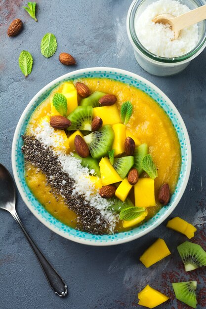 Ciotola per frullato di mango con pezzi di kiwi, mango, scaglie di cocco e semi di chia su una pietra