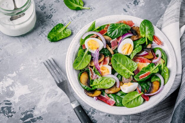 Ciotola per il pranzo di insalata di spinaci con uova di funghi bacon e cipolle rosse su sfondo grigio