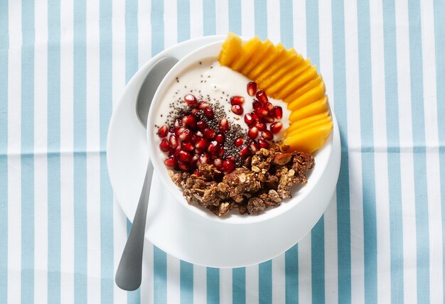 Ciotola per la colazione di muesli, yogurt, mango dolce fresco, semi di chia, semi di melograno e sciroppo d'acero su un tovagliolo a strisce blu