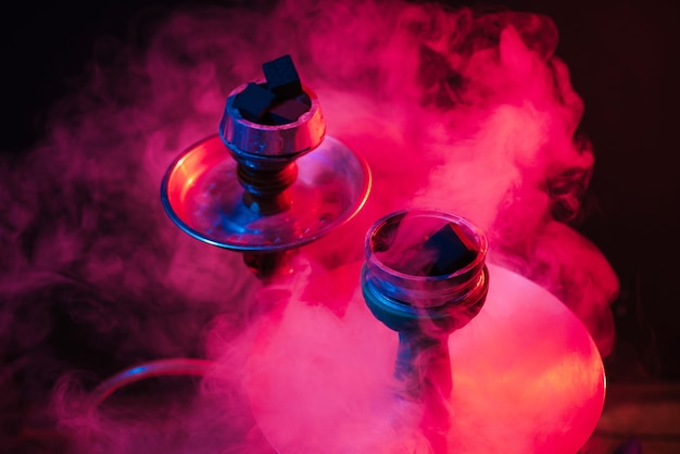 Ciotola per narghilè, narghilè e carboni ravvicinati su uno sfondo nero fumoso con illuminazione colorata