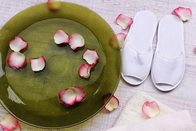 Ciotola Spa con acqua, petali di rosa, asciugamano e pantofole su sfondo chiaro. Concetto di pedicure o trattamento termale naturale