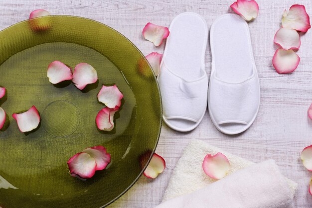 Ciotola Spa con acqua, petali di rosa, asciugamano e pantofole su sfondo chiaro. Concetto di pedicure o trattamento termale naturale