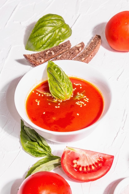 Ciotole con zuppa di pomodoro fatta in casa. Verdure mature, foglie di basilico fresco, grissini, spezie aromatiche. Luce dura alla moda, ombra scura. Sfondo bianco stucco, distesi piatti