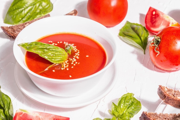 Ciotole con zuppa di pomodoro fatta in casa. Verdure mature, foglie di basilico fresco, grissini, spezie aromatiche. Luce dura alla moda, ombra scura. Sfondo bianco stucco, distesi piatti