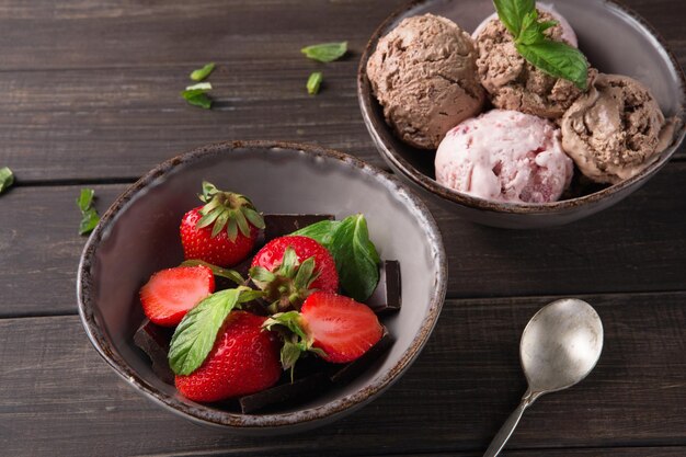 Ciotole di cioccolato e gelato alla bacca e fragole su fondo di legno rustico con foglie di menta e cucchiaio, primo piano. Dolce estivo fatto in casa