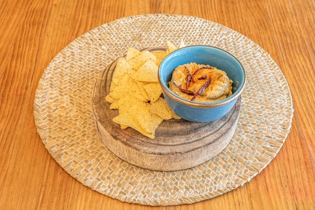 Ciotolina blu con hummus di ceci scaglie di peperone essiccato e nachos da intingere su un pezzo di tronco