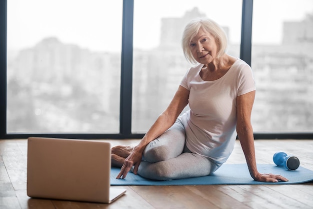 Classe in linea. Donna anziana dai capelli grigi seduta sul pavimento e che tiene una lezione di yoga online