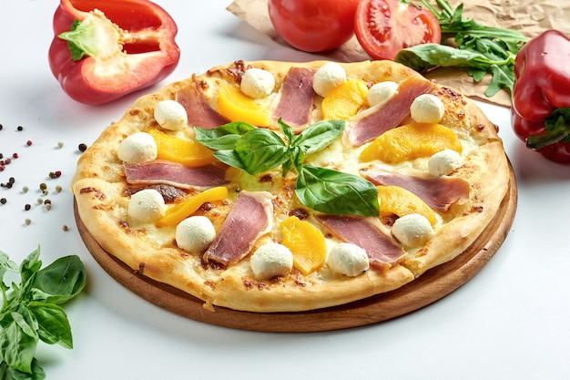 Classica pizza italiana al prosciutto, crema di formaggio, pesche e basilico su un piatto. Superficie bianca