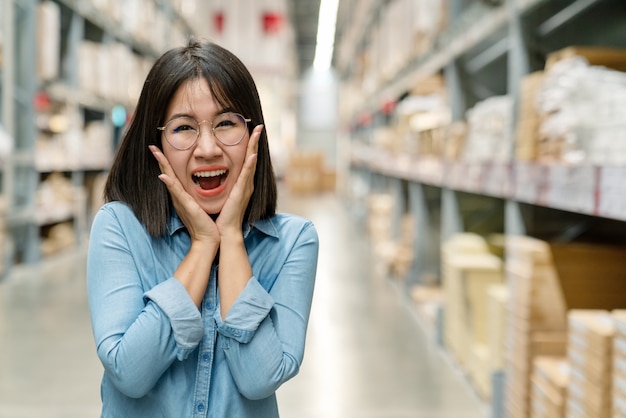 Cliente asiatico sorpreso della donna che guarda macchina fotografica e che ritiene soddisfatto dello sconto e della vendita. Gioia e divertimento asiatici dell'imprenditore con controllo dell'inventario nel magazzino.