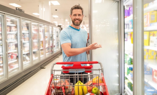 Cliente consumatore con acquisti ragazzo al negozio di alimentari acquirente a generi alimentari che acquista cibo al supermercato consumo che presenta prodotto uomo allegro con carrello