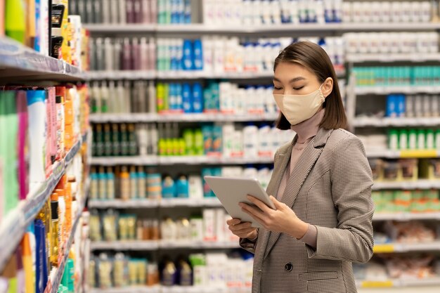 Cliente femminile matura contemporanea che scorre l'elenco dei prodotti alimentari da acquistare nel supermercato mentre passa dall'esposizione di frutta