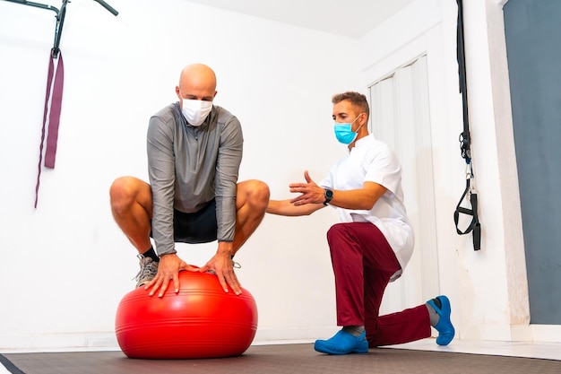 Cliente in equilibrio su pallone rosso assistito da Fisioterapista con mascherina Fisioterapia con misure protettive per la pandemia di Coronavirus COVID19 Osteopatia sport quiromassaggio