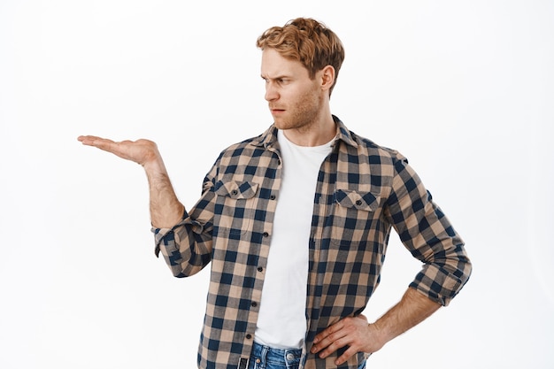 Cliente maschio deluso arrabbiato che guarda la sua mano aperta che tiene l'oggetto, non gli piace un oggetto che tiene sul palmo, fissando scontento, in piedi sul muro bianco