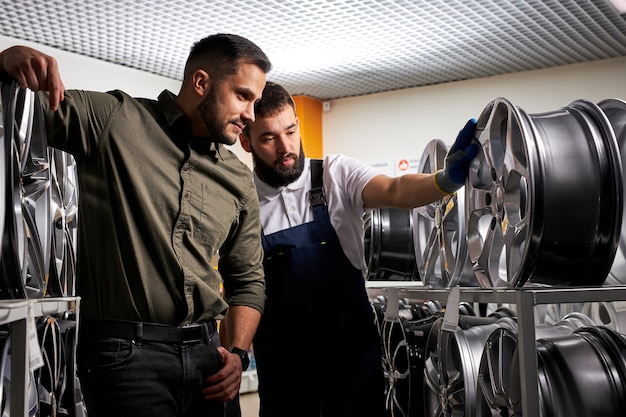 Cliente piacevole e meccanico auto fiducioso che parla di ruote di auto, in negozio, in piedi tra cremagliera di cerchi