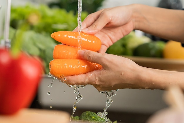 Close up asiatico giovane donna lavaggio carota pomodoro broccoli verdure fresche paprika con spruzzi d'acqua nella bacinella d'acqua sul lavandino in cucina preparare insalata fresca cucinare pasto sano cibo persone