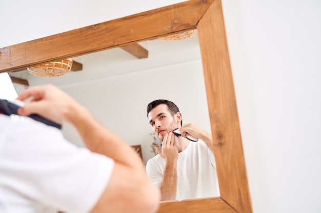 Close up brunet uomo in maglietta bianca si rade mentre si trova vicino allo specchio nella vasca da bagno al mattino
