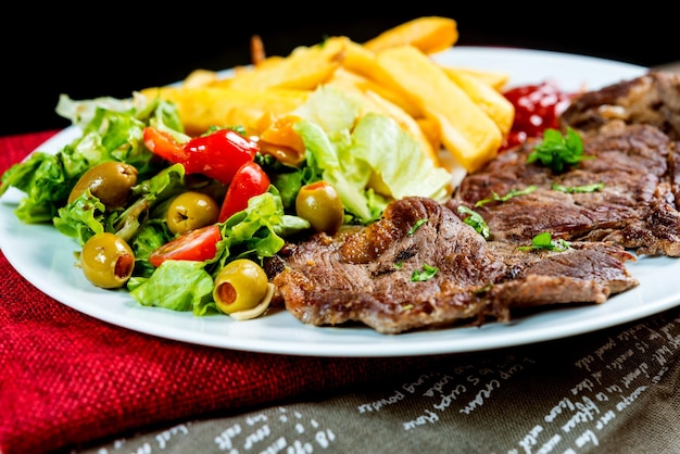 Close-up di bistecca di manzo con insalata e patatine fritte sul tavolo contro uno sfondo nero