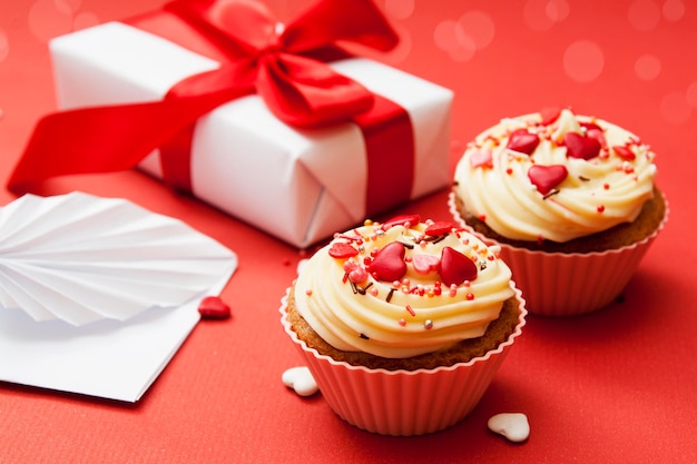 Close-up di due cupcakes con crema e decorazioni a cuore su sfondo rosso con regalo e busta. Composizione per San Valentino.