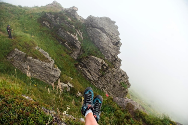 Close up di gambe maschili in scarpe da ginnastica sportive sedute sul bordo delle montagne tra la natura verde Donna in escursione con pali da passeggio in distanza Concetto di turismo