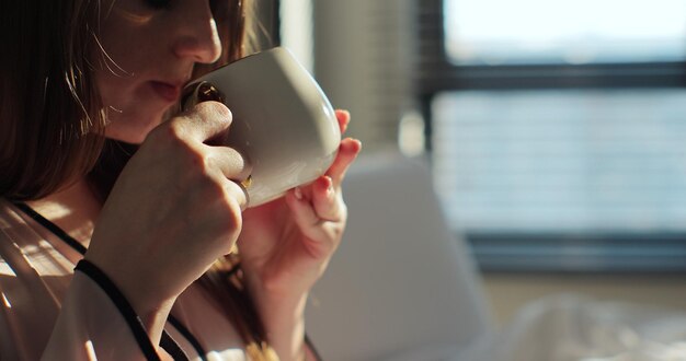 Close up di una giovane donna attraente che beve una tazza di caffè o tè nel letto e si diverte