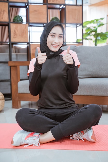 Close up musulmano donna che indossa un hijab allenamento vestito si siede a gambe incrociate sul pavimento con un pollice in alto nel soggiorno