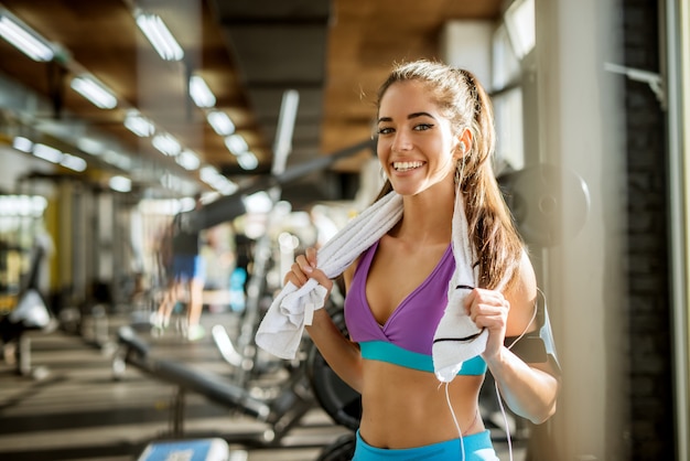 Close up ritratto di adorabile affascinante sorridente giovane forma fitness ragazza con gli auricolari in possesso di un asciugamano e in posa mentre guarda la telecamera in palestra vicino alla finestra.