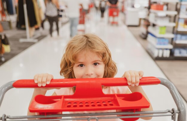 Close up ritratto di bambino con carrello acquisto di cibo in un negozio di alimentari. Famiglia di clienti che acquistano prodotti al supermercato. Viso divertente per bambini.