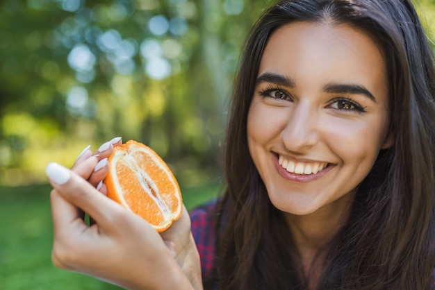 Close up ritratto di bella bella bruna donna sorridente che tiene succosa deliziosa arancia su sfondo verde parco Giovane donna con un sorriso sano che tiene frutta Copia spazio per il contenuto pubblicitario