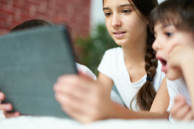 Close up ritratto di bella ragazza latina sorridente durante l'utilizzo di tablet digitale. Sorella adolescente che intrattiene i suoi due fratellini, guardando i cartoni animati a casa. Infanzia felice, concetto di tecnologia